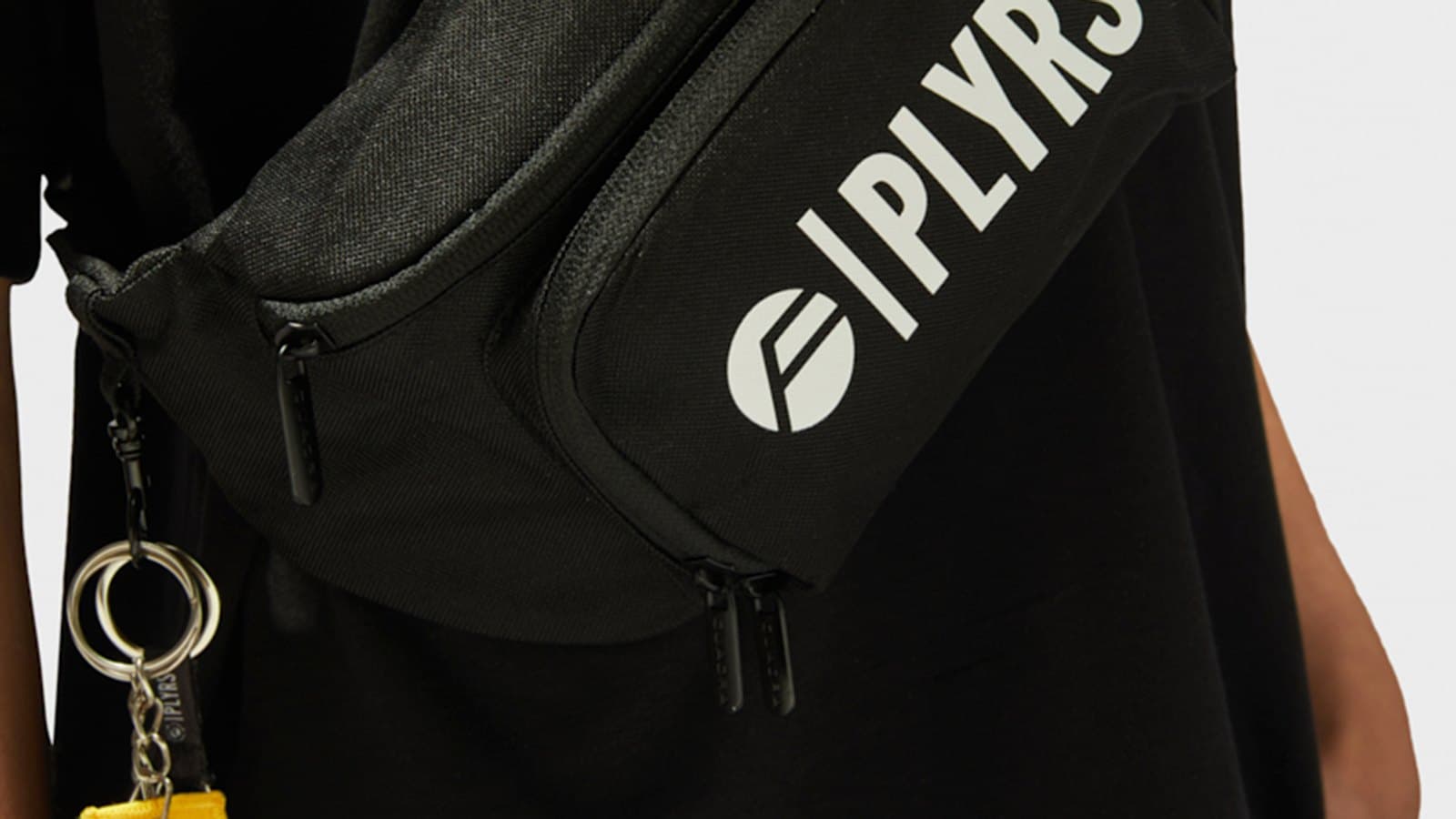 Close-up of a PLYRS fanny pack and keychain on a black coach jacket and joggers